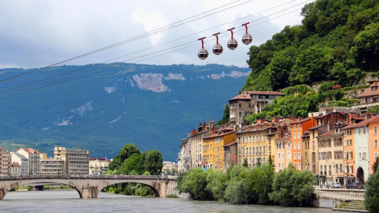 Une rangée de bâtiments colorés borde une rivière, traversée par un pont de pierre. Quatre téléphériques sphériques glissent au-dessus, entourés de collines verdoyantes et de montagnes escarpées sous un ciel partiellement nuageux.
