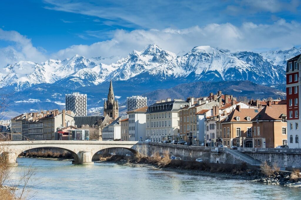 Un paysage urbain pittoresque composé de bâtiments colorés, d'un pont de pierre enjambant une rivière et d'une église avec une haute flèche, sur fond de montagnes enneigées sous un ciel bleu.
