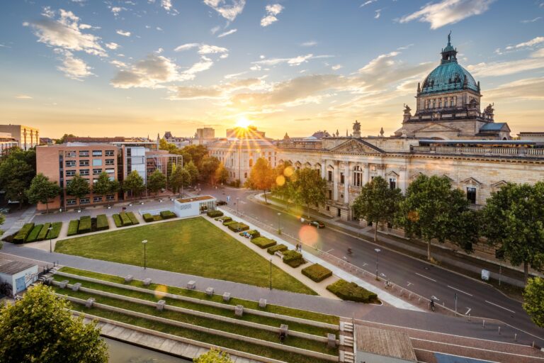 Un paysage urbain au coucher du soleil présente un bâtiment historique en forme de dôme à côté d'un grand parc verdoyant et de structures modernes. La lumière du soleil traverse les nuages et jette une lueur chaude sur les arbres et les rues vides.