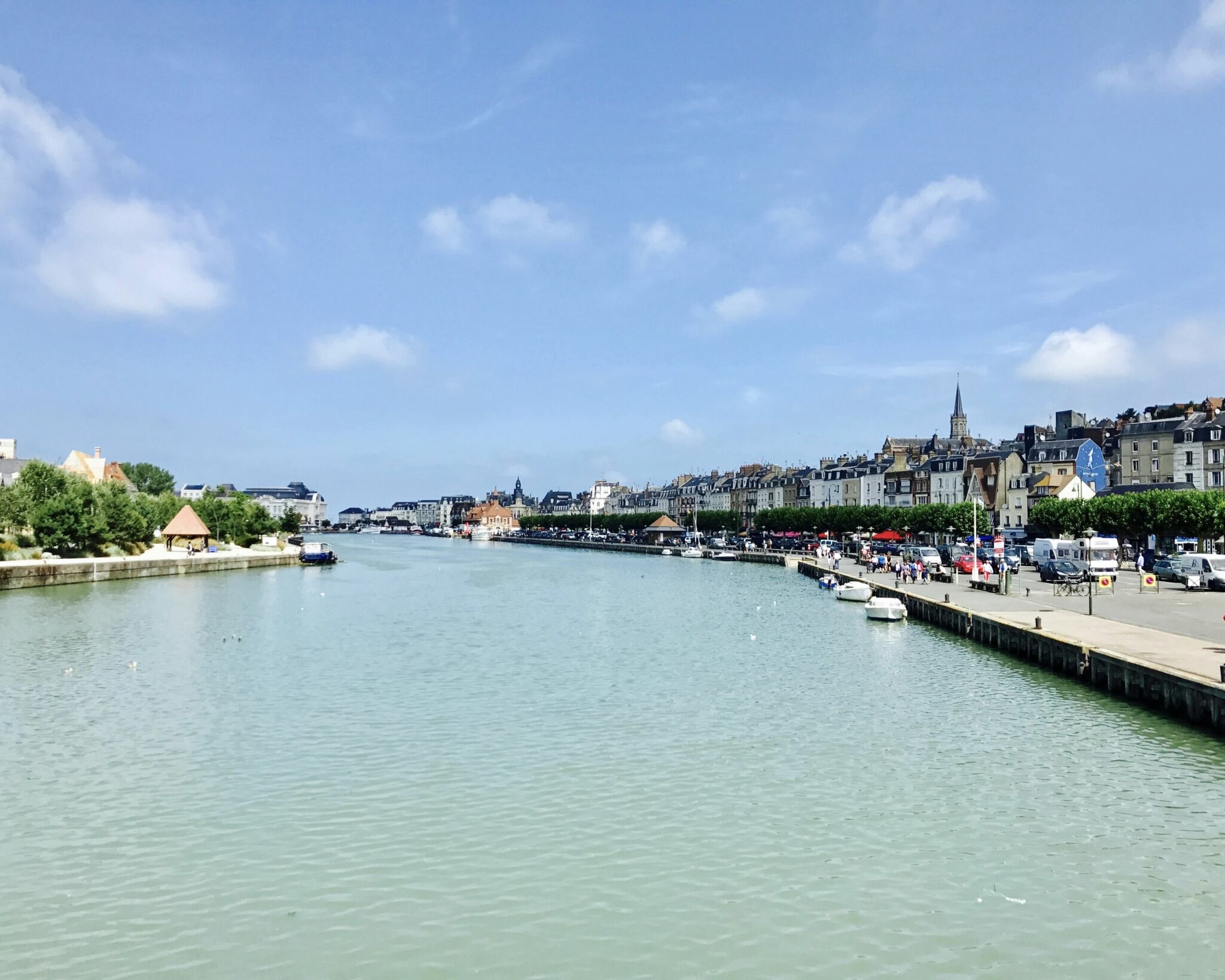 Une large rivière bordée de bateaux traverse une ville avec des bâtiments historiques et une flèche d'église. Des arbres et une promenade longent la rive droite sous un ciel lumineux et partiellement nuageux.