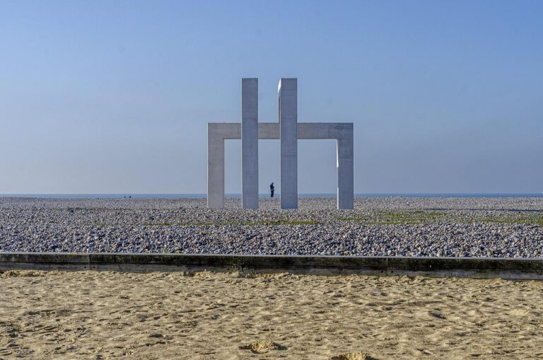 Une grande sculpture géométrique en béton se dresse sur une plage rocheuse sous un ciel bleu clair, avec une seule personne visible au loin à travers la structure. Le sable est au premier plan.