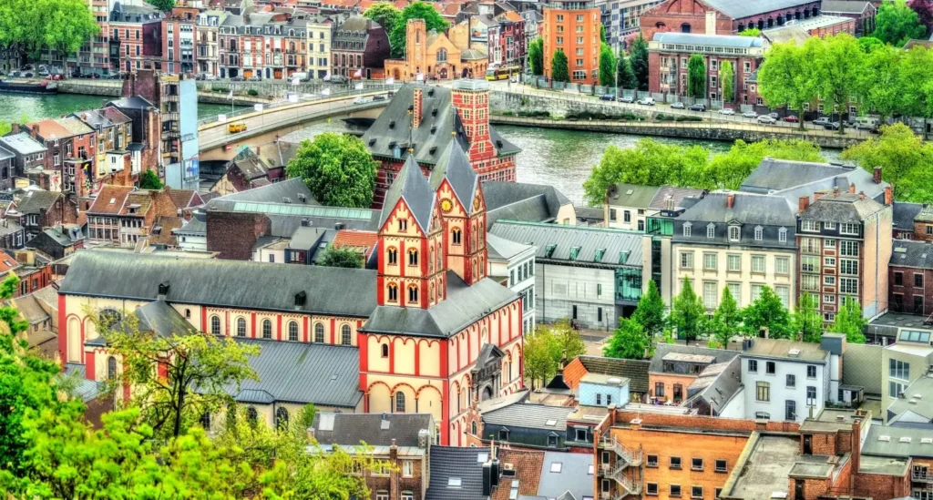 Des bâtiments historiques colorés entourent une grande église aux accents rouges et blancs dans une ville européenne au bord d'une rivière, avec un pont et des arbres verts en arrière-plan.