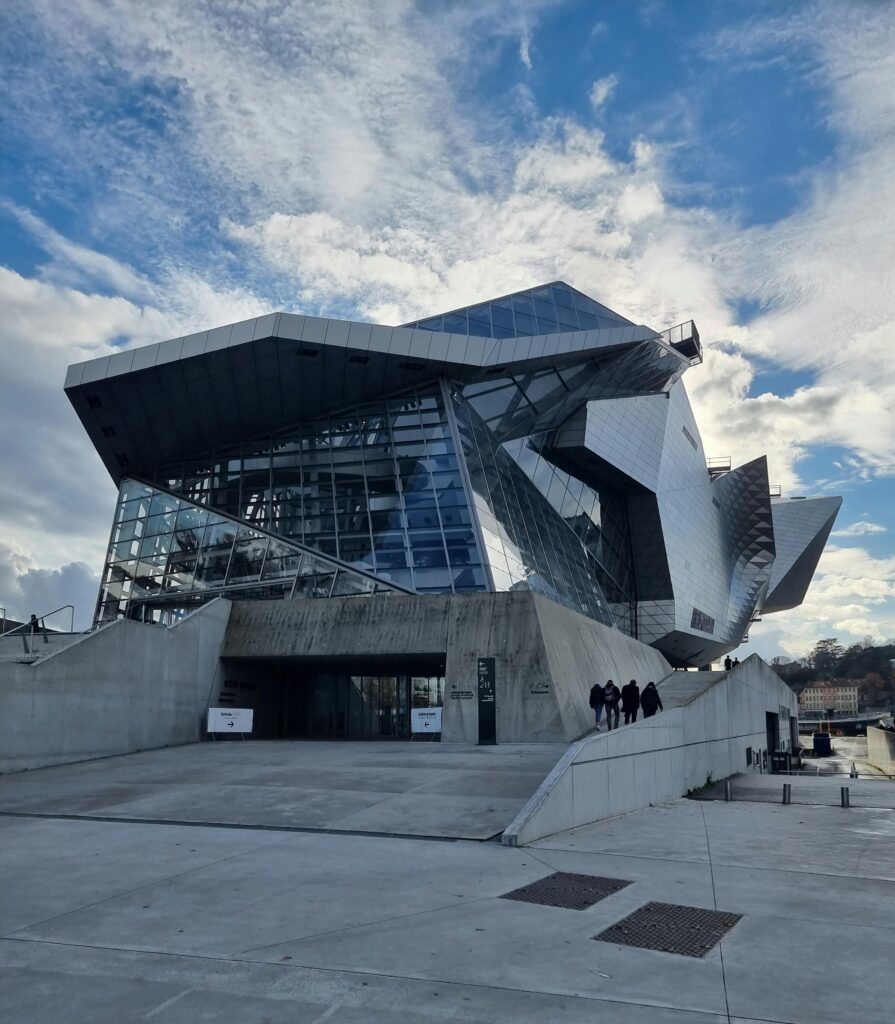 Un bâtiment moderne et anguleux avec des façades en verre et en métal sous un ciel partiellement nuageux. Plusieurs personnes se dirigent vers l'entrée en empruntant une large rampe en béton.