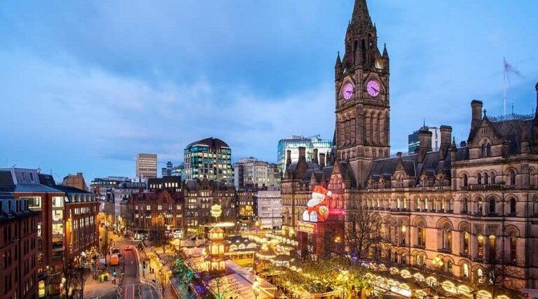 Une scène de ville festive au crépuscule montre l'hôtel de ville de Manchester illuminé avec sa tour d'horloge, entouré de lumières de Noël, d'étals de marché et d'une foule animée dans le centre ville.