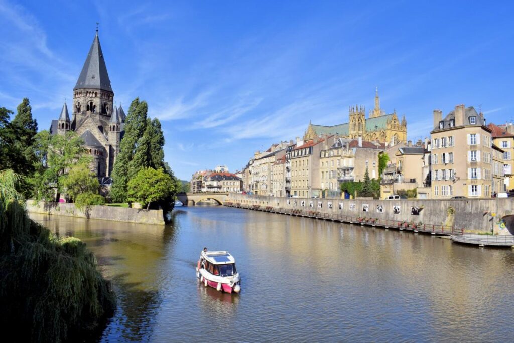 A small boat sails along a calm river lined with historic buildings and a church with a high spire, under a clear blue sky. Trees line the bank and a stone bridge can be seen in the background.