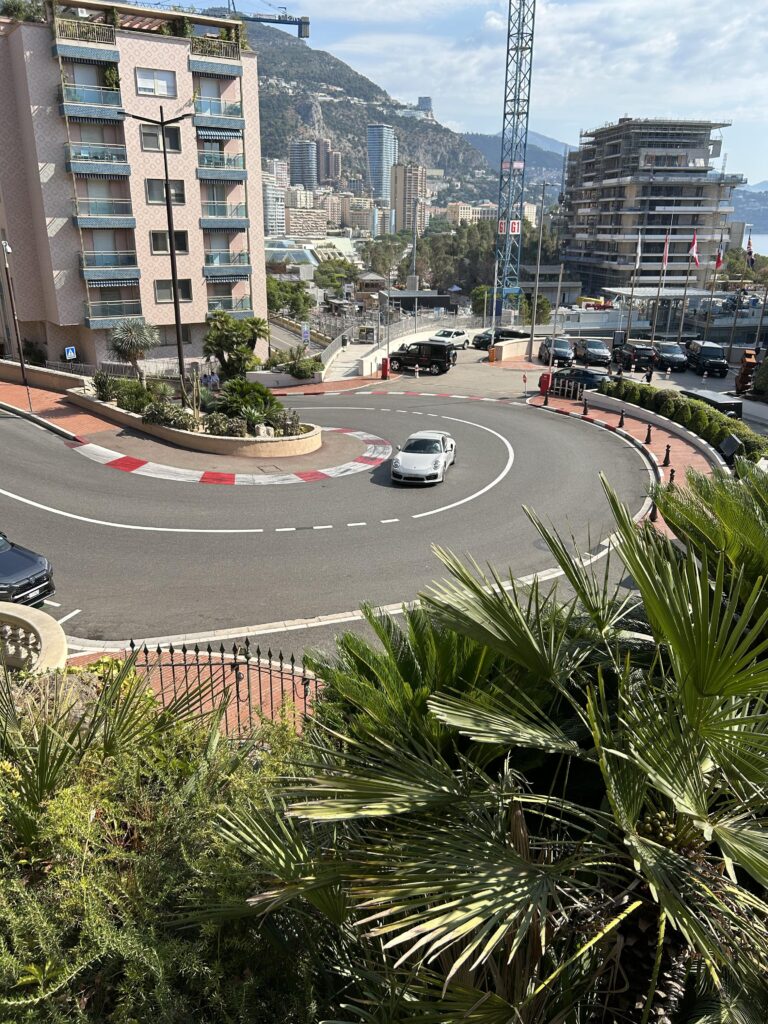 Une voiture de sport blanche négocie un virage serré dans une rue de la ville entourée d'immeubles et de palmiers, avec un arrière-plan montagneux et un ciel partiellement nuageux.