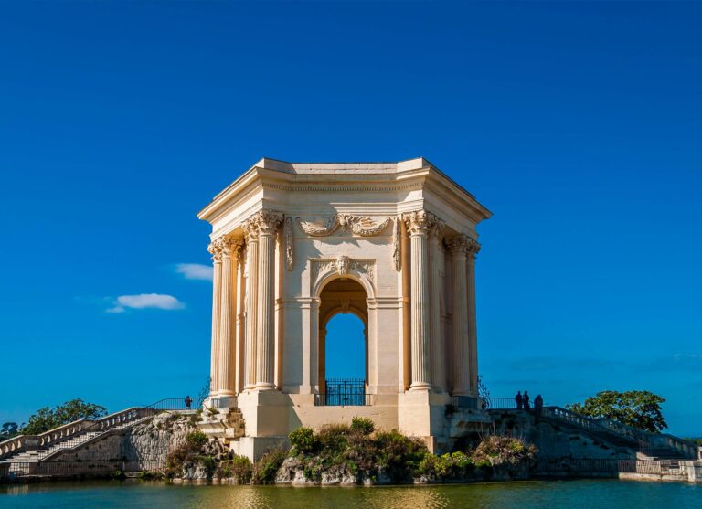 Un pavillon néoclassique en pierre avec des colonnes se trouve à côté d'un étang sous un ciel bleu clair, avec quelques personnes visibles près de la structure et des arbres à l'arrière-plan.