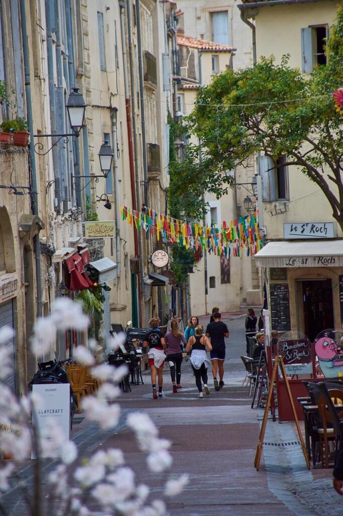 Une rue européenne étroite bordée de vieux bâtiments, de tables de café en plein air et de drapeaux colorés suspendus au-dessus. Cinq personnes marchent sur le chemin pavé, tandis que des fleurs apparaissent au premier plan.