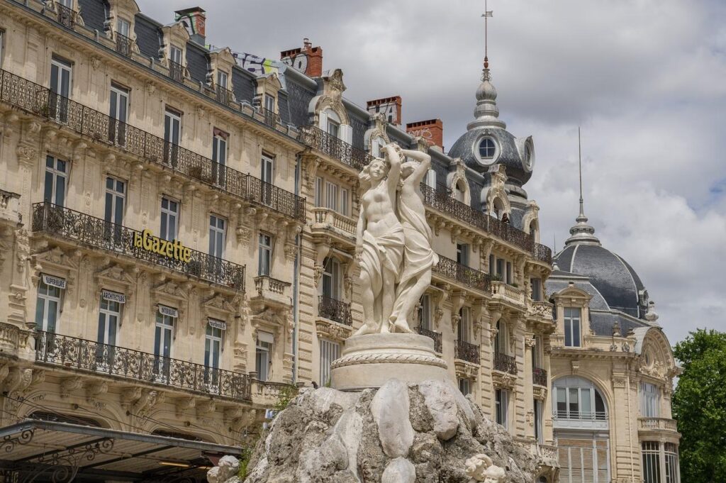 Une statue de pierre blanche représentant deux personnages se dresse sur un socle rocheux devant des bâtiments historiques ornés de balcons en fer forgé et de toits en dôme, sous un ciel nuageux.
