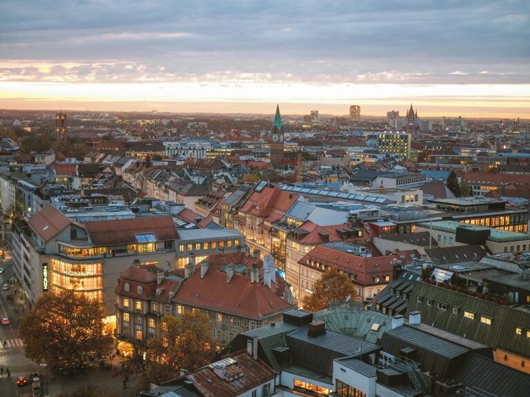 Vue aérienne d'une ville au coucher du soleil avec un mélange de bâtiments modernes et historiques, des toits rouges, des lampadaires lumineux et une église avec un clocher vert au centre.