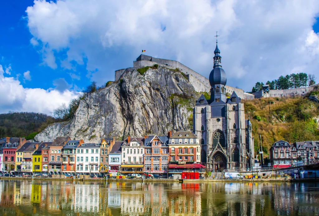Des bâtiments colorés bordent le front de mer à Dinant, en Belgique, avec la collégiale ornée de Notre-Dame et une forteresse perchée sur une falaise rocheuse, le tout se reflétant dans la rivière calme sous un ciel partiellement nuageux.