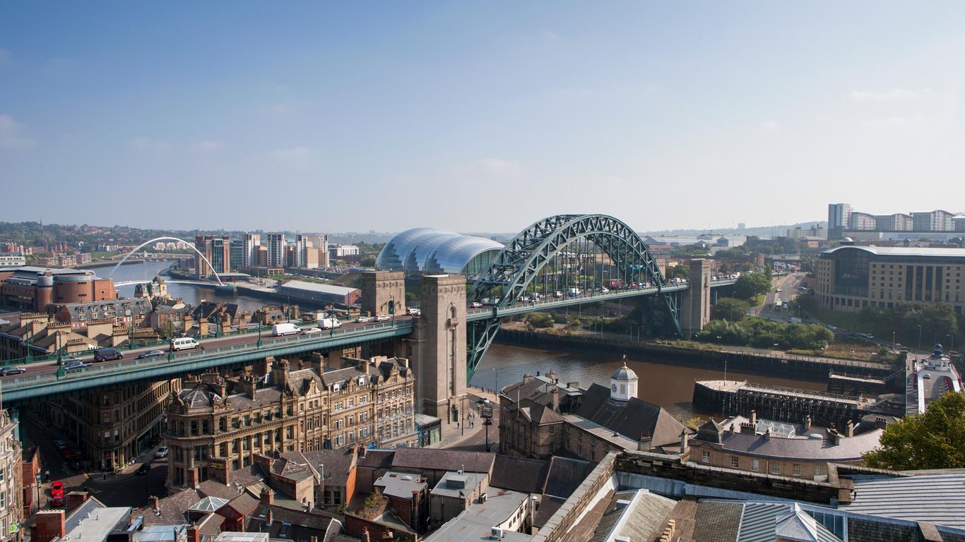 Vue panoramique de Newcastle upon Tyne avec le Tyne Bridge qui enjambe la rivière, des bâtiments historiques au premier plan et la salle de concert moderne Sage Gateshead à l'arrière-plan, sous un ciel bleu clair.
