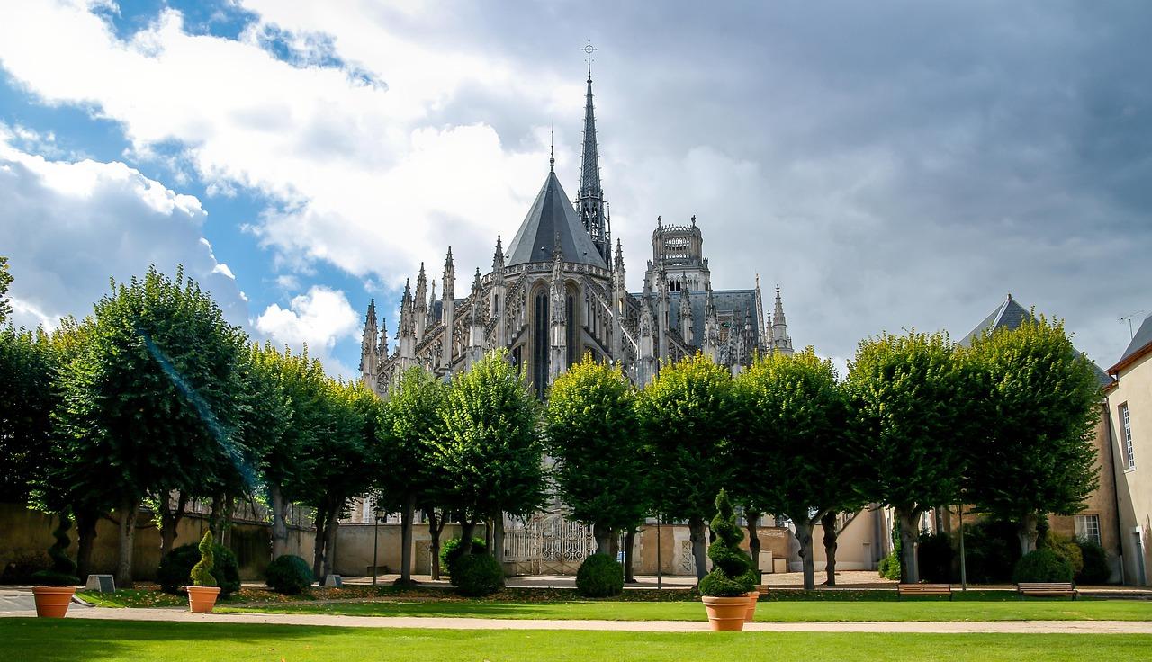 Une cathédrale gothique avec de hautes flèches s'élève derrière une rangée d'arbres verts dans un jardin manucuré, sous un ciel partiellement nuageux.