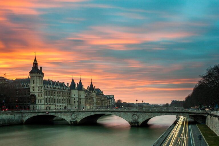 Vue panoramique de la Conciergerie et du pont au Change sur la Seine à Paris au coucher du soleil, avec des nuages colorés dans le ciel et des traînées lumineuses provenant des véhicules qui passent le long de la route riveraine.