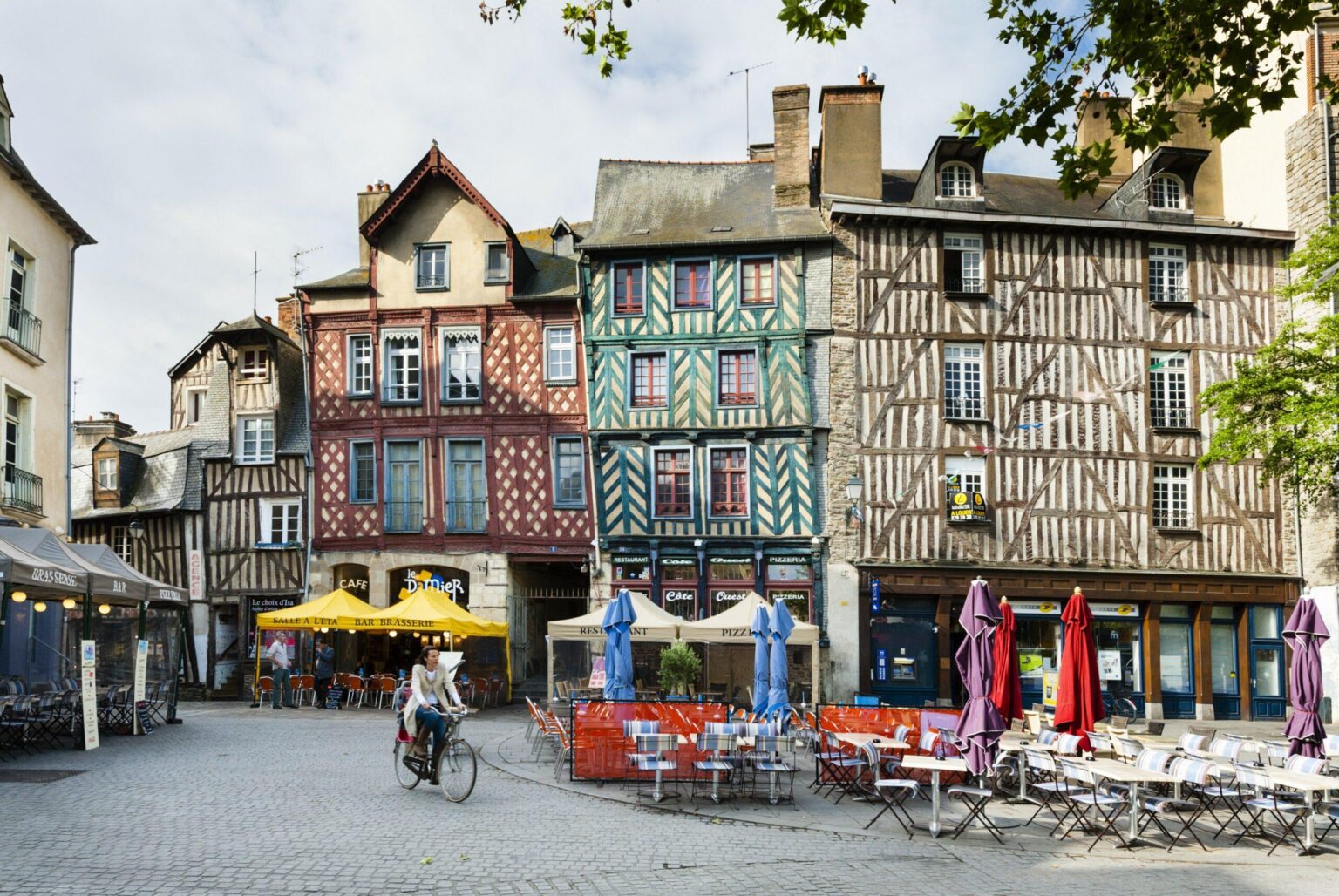 Un cycliste traverse une place pavée bordée de bâtiments à colombages colorés, de cafés en plein air et de tables vides avec des parapluies fermés dans une ville européenne historique.
