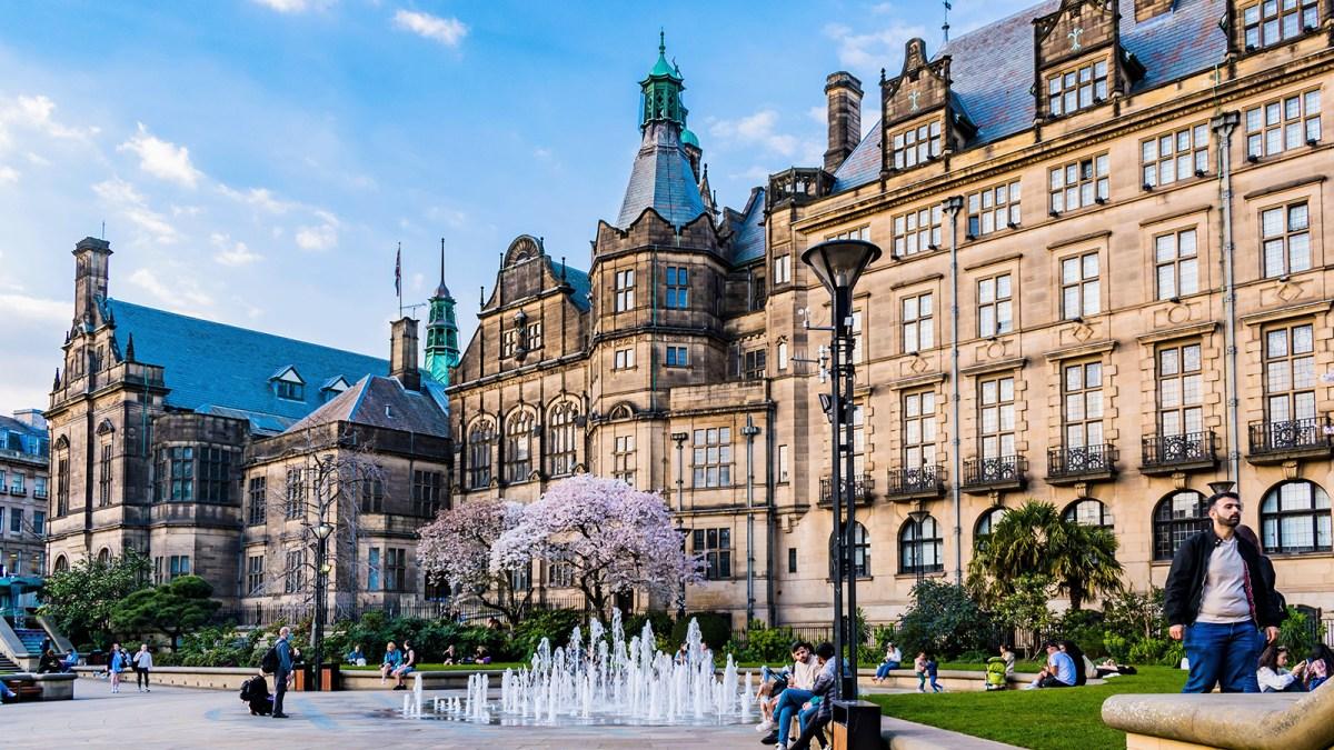 Des personnes se détendent près d'une petite fontaine et d'un cerisier en fleurs sur une place de la ville entourée d'un bâtiment historique orné, sous un ciel bleu.