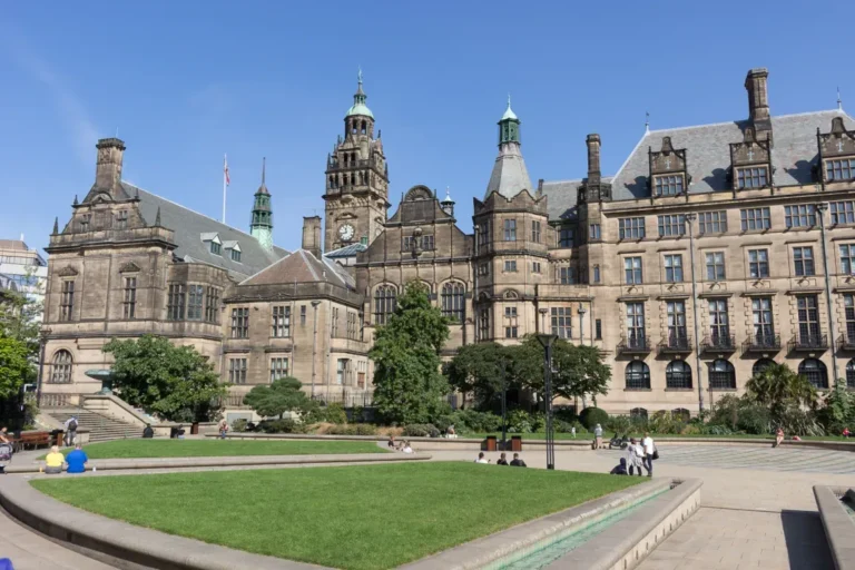 Un grand bâtiment historique avec une architecture en pierre ornée, des tours et une tour d'horloge, entouré d'une cour paysagée avec des pelouses vertes, des arbres et des gens qui se détendent sous un ciel bleu clair.