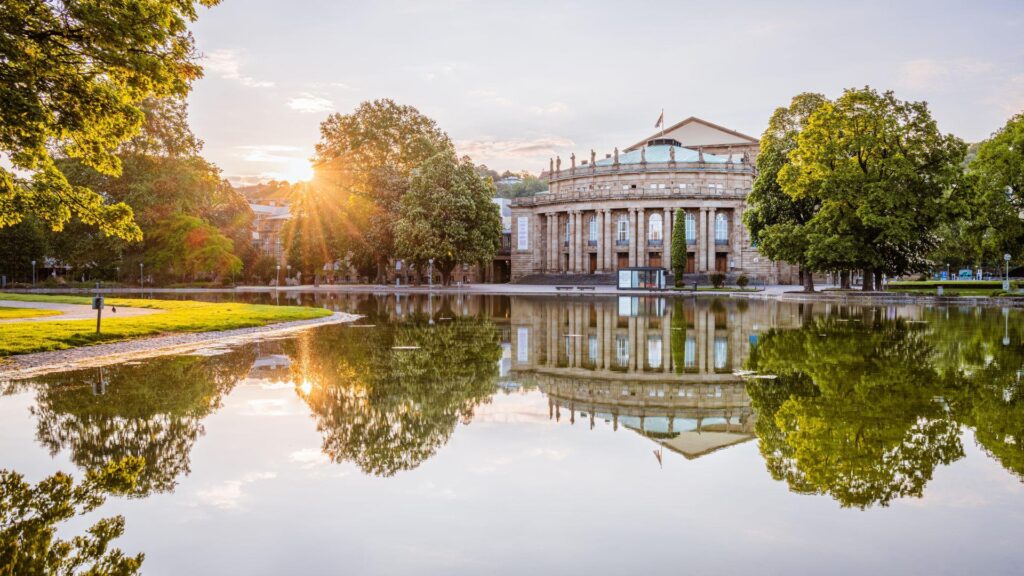 Le soleil se couche derrière un grand bâtiment néoclassique à colonnes, se reflétant dans un étang calme entouré d'arbres verts dans un parc.