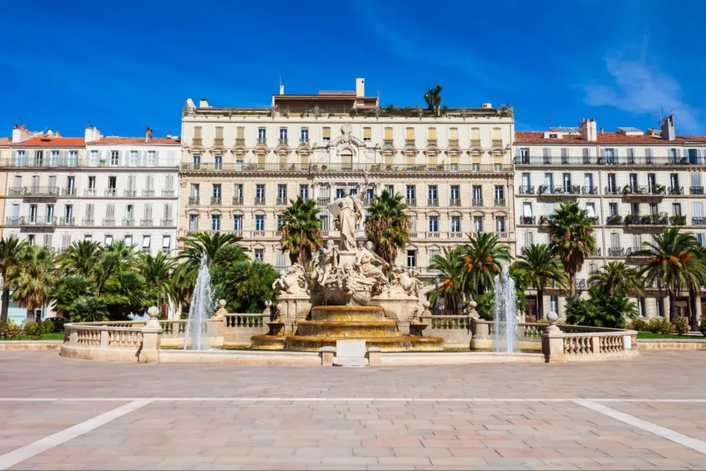 Une grande fontaine ornée de statues se dresse devant des palmiers et plusieurs bâtiments hauts et élégants, sous un ciel bleu clair, sur une vaste place de la ville.