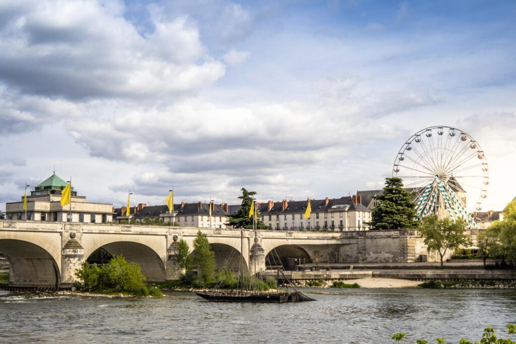 Un pont de pierre enjambe une rivière avec des bâtiments et des arbres en arrière-plan ; une grande roue s'élève au-dessus des toits sous un ciel partiellement nuageux. Des drapeaux jaunes ornent le pont.