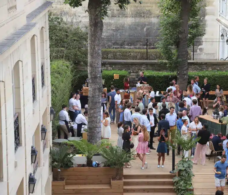 Un groupe de personnes se retrouve lors d'un événement en plein air, dans un cadre agrémenté de tables, de plantes et d'arbres. Certains invités discutent tandis que d'autres prennent des photos ou sont assis. L'ambiance qui règne dans cette cour est festive et détendue. Un groupe de personnes se retrouve lors d'un événement en plein air, dans un cadre agrémenté de tables, de plantes et d'arbres. Certains invités discutent tandis que d'autres prennent des photos ou sont assis. L'ambiance qui règne dans cette cour est festive et détendue.
