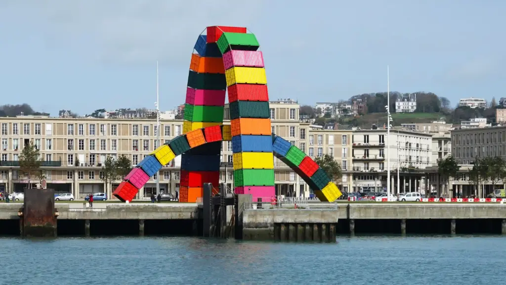 Des conteneurs d'expédition aux couleurs vives sont empilés en forme d'arche sur un quai au bord de l'eau, avec en arrière-plan les bâtiments de la ville et une colline. Des conteneurs d'expédition aux couleurs vives sont empilés en forme d'arche sur un quai au bord de l'eau, avec en arrière-plan les bâtiments de la ville et une colline.