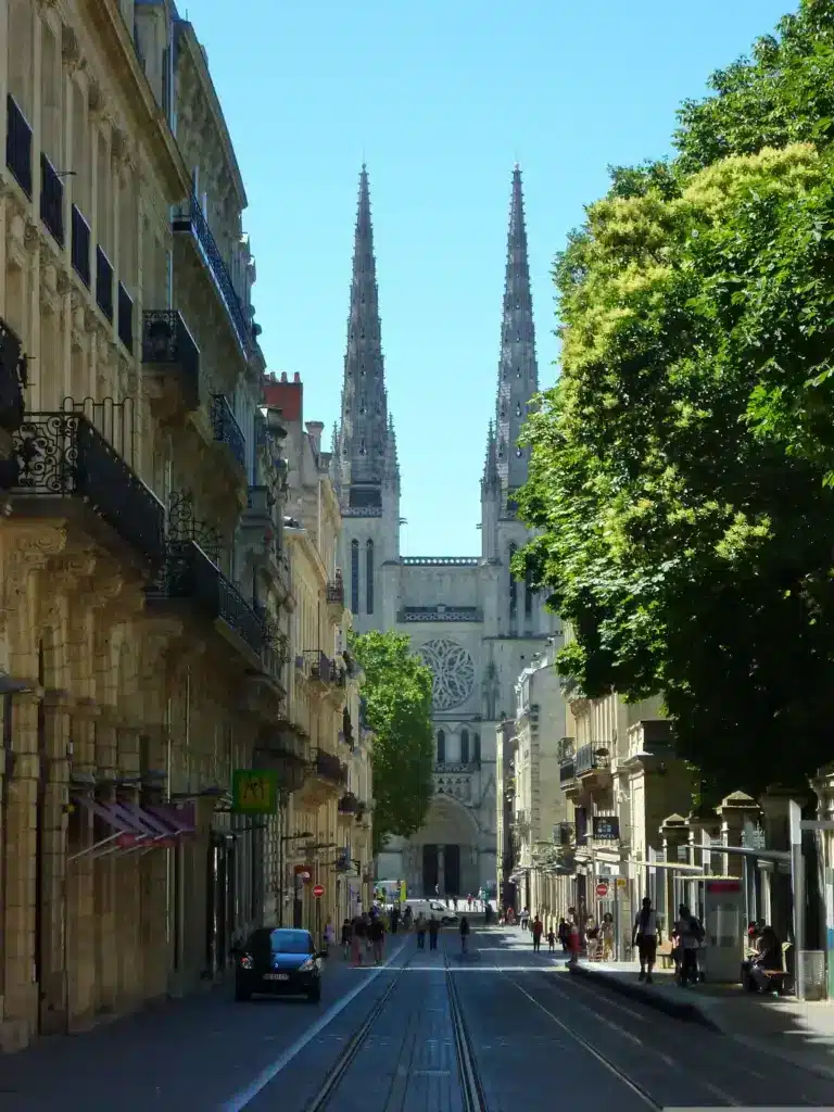 Une rue de la ville, bordée de rails de tramway, mène à une grande cathédrale dotée de deux hautes flèches. Des bâtiments historiques et des arbres verdoyants bordent la rue sous un ciel bleu limpide. Des gens se promènent et quelques voitures sont garées. Une rue de la ville, bordée de rails de tramway, mène à une grande cathédrale dotée de deux hautes flèches. Des bâtiments historiques et des arbres verdoyants bordent la rue sous un ciel bleu limpide. Des gens se promènent et quelques voitures sont garées.