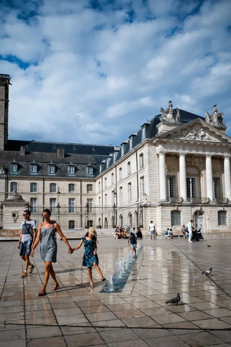Des gens traversent une place mouillée parsemée de petites fontaines. En arrière-plan, sous un ciel partiellement nuageux, on aperçoit des bâtiments de style classique ornés de colonnes et de statues. Des pigeons sont visibles au sol.