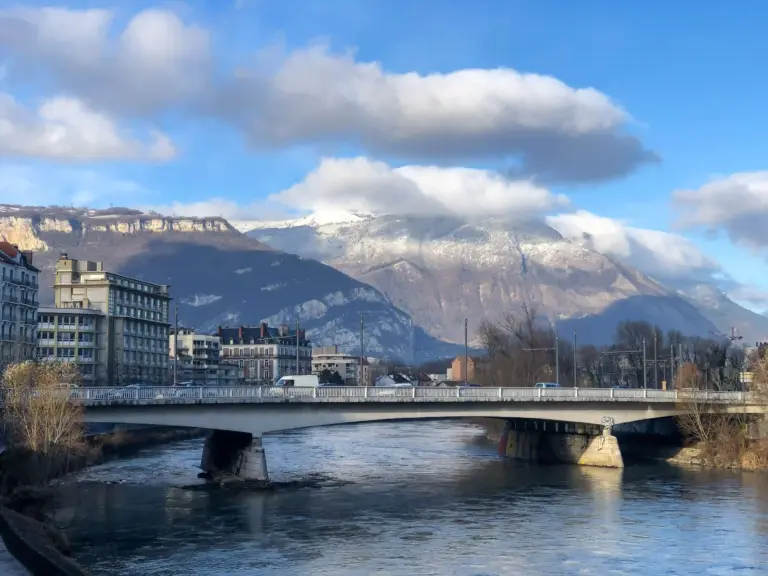 Un pont en béton enjambe une rivière paisible, avec des bâtiments sur la gauche et des montagnes enneigées en arrière-plan, sous un ciel bleu partiellement nuageux. Un pont en béton enjambe une rivière paisible, avec des bâtiments sur la gauche et des montagnes enneigées en arrière-plan, sous un ciel bleu partiellement nuageux.