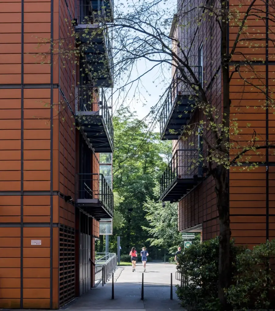 Deux personnes marchent entre deux immeubles modernes de couleur orange dotés de balcons métalliques ; on aperçoit des arbres en arrière-plan, et la lumière du soleil inonde cet espace ouvert.
