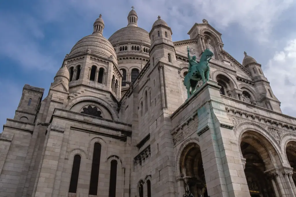 La basilique du Sacré-Cœur à Paris, vue d'en bas, avec ses dômes blancs et une statue équestre verte devant, sur fond de ciel partiellement nuageux.