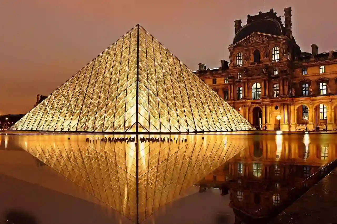 L'entrée en forme de pyramide de verre illuminée du musée du Louvre à Paris, la nuit, se reflétant dans l'eau calme, avec le bâtiment historique du musée visible à côté.