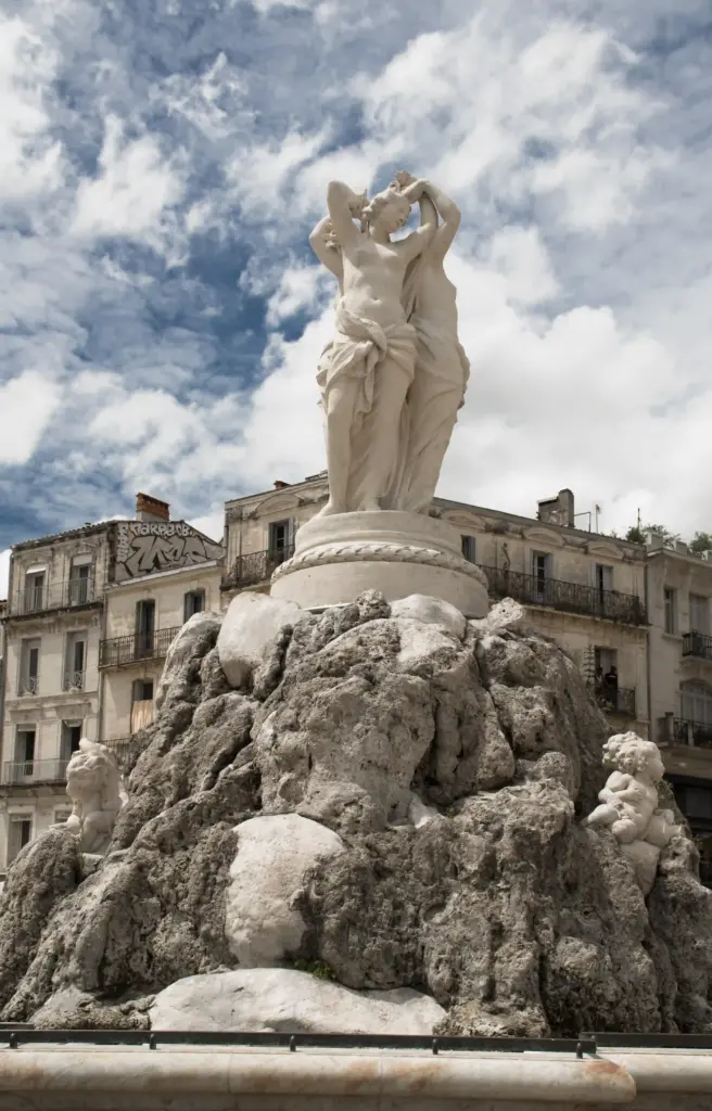 Une statue en marbre blanc représentant deux femmes trône au sommet d'un socle de fontaine rocheux, entourée de bâtiments anciens sous un ciel partiellement nuageux. Une statue en marbre blanc représentant deux femmes trône au sommet d'un socle de fontaine rocheux, entourée de bâtiments anciens sous un ciel partiellement nuageux.