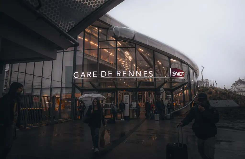 Des passants marchent devant la Gare de Rennes, une gare moderne aux façades vitrées située à Rennes, en France, par un jour de pluie. Le logo de la SNCF est visible au-dessus de l'entrée. Certaines personnes portent des bagages et des parapluies. Des passants marchent devant la Gare de Rennes, une gare moderne aux façades vitrées située à Rennes, en France, par un jour de pluie. Le logo de la SNCF est visible au-dessus de l'entrée. Certaines personnes portent des bagages et des parapluies.