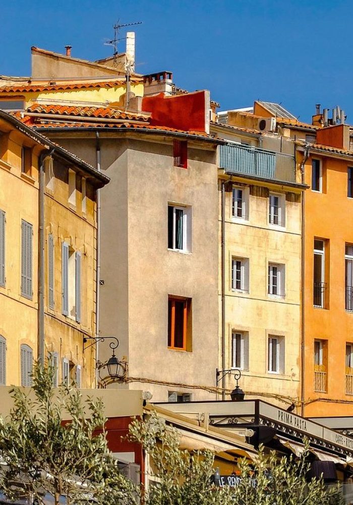 Rangée colorée et ensoleillée de bâtiments hauts et étroits avec des volets et des balcons dans une ville européenne, avec des cafés avec des sièges en plein air et des auvents au niveau de la rue sous un ciel bleu clair.