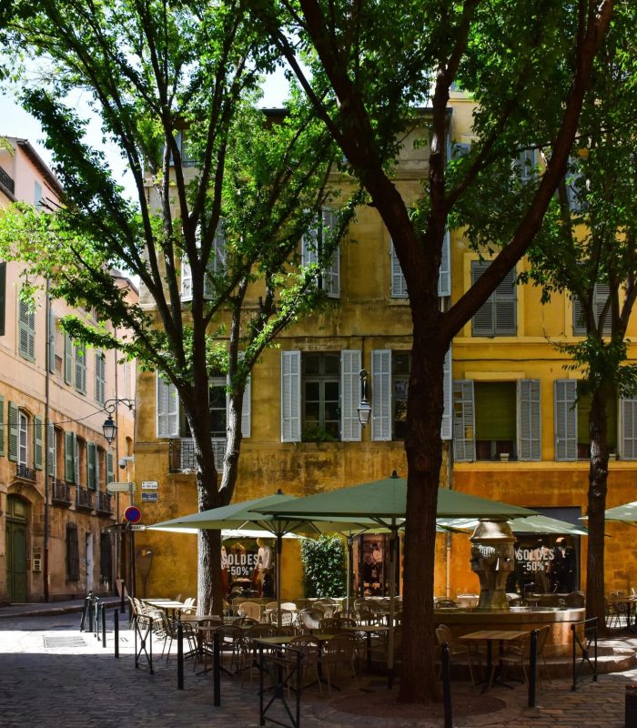 Une rue européenne ensoleillée avec des bâtiments historiques, de grands arbres verts et un café en plein air avec des tables et des parasols. Deux personnes marchent sur la route pavée et le quartier semble calme et accueillant.