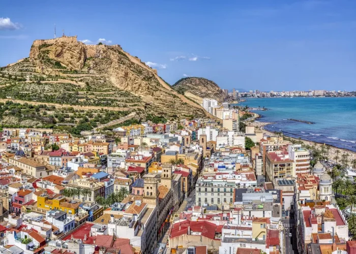 Bâtiments colorés et paysage urbain d'Alicante, Espagne, avec le château de Santa Bárbara sur une colline rocheuse surplombant la ville et la mer Méditerranée sous un ciel bleu clair.