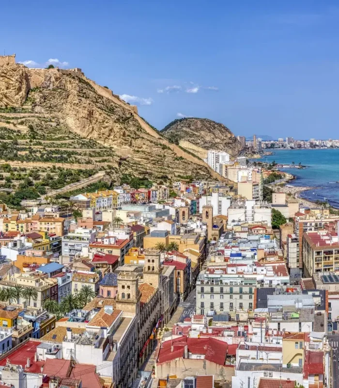Bâtiments colorés et paysage urbain d'Alicante, Espagne, avec le château de Santa Bárbara sur une colline rocheuse surplombant la ville et la mer Méditerranée sous un ciel bleu clair.
