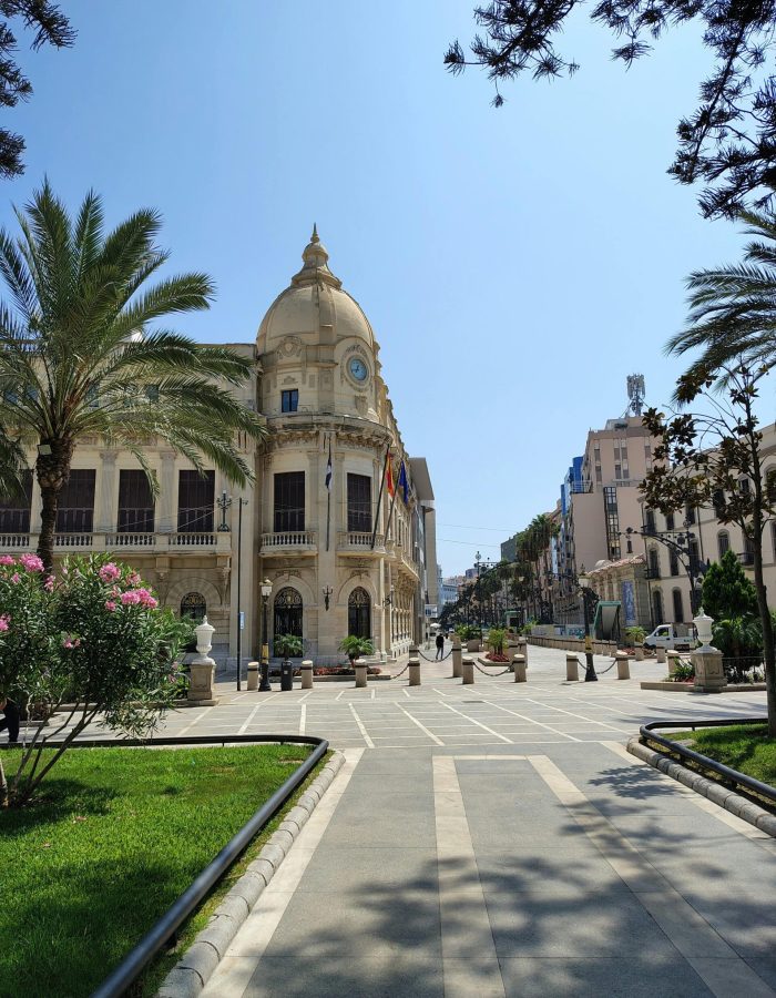 Un grand bâtiment historique avec une tour d'horloge en forme de dôme se dresse à l'extrémité d'une place ensoleillée bordée de palmiers, de buissons fleuris et de pelouses manucurées sous un ciel bleu clair.