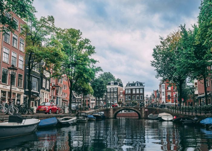 Un canal d'Amsterdam bordé de bateaux, de vélos et d'arbres, bordé de bâtiments hauts et étroits, avec un pont en pierre traversant l'eau sous un ciel nuageux.