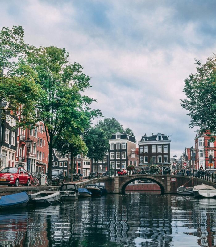 Un canal d'Amsterdam bordé de bateaux, de vélos et d'arbres, bordé de bâtiments hauts et étroits, avec un pont en pierre traversant l'eau sous un ciel nuageux.