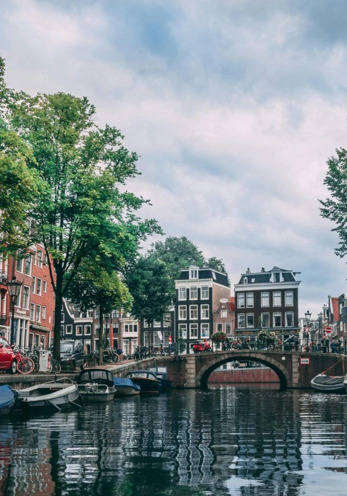 Un canal d'Amsterdam bordé de bateaux, de vélos et d'arbres, bordé de bâtiments hauts et étroits, avec un pont en pierre traversant l'eau sous un ciel nuageux.