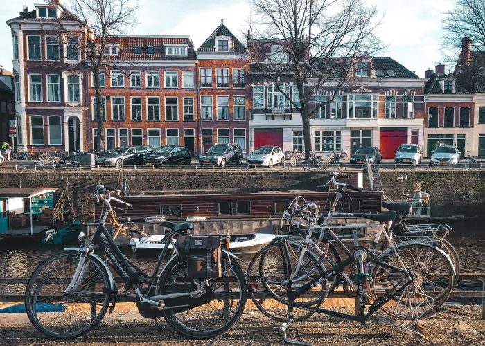 Plusieurs vélos sont garés près d'un canal avec de l'eau, et des rangées de bâtiments européens traditionnels avec de grandes fenêtres et des façades rouges et blanches sont visibles à l'arrière-plan. Des arbres sans feuilles bordent la rue.