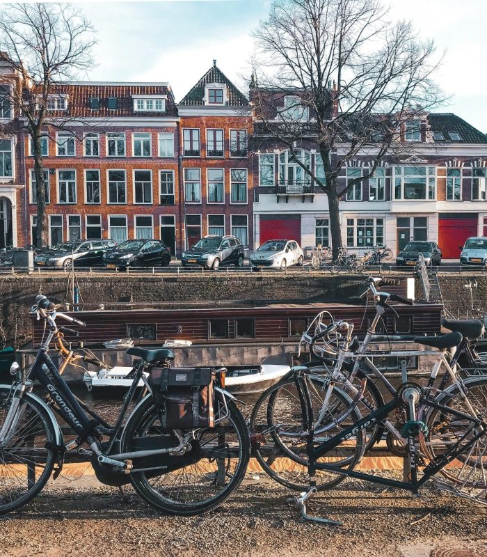 Plusieurs vélos sont garés près d'un canal avec de l'eau, et des rangées de bâtiments européens traditionnels avec de grandes fenêtres et des façades rouges et blanches sont visibles à l'arrière-plan. Des arbres sans feuilles bordent la rue.