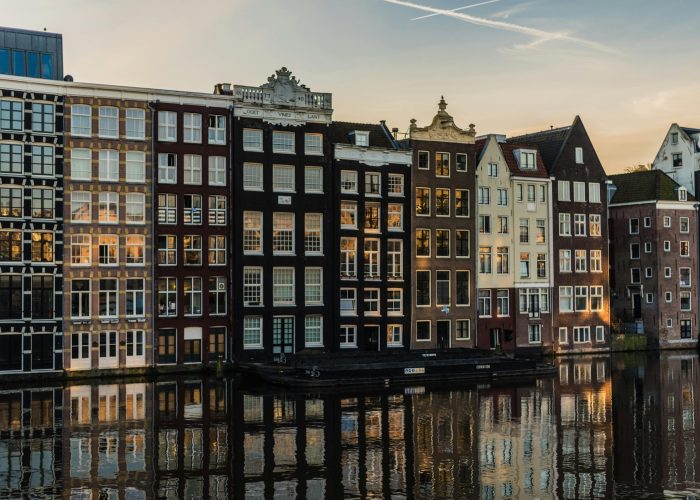 Rangée de maisons hautes et étroites avec de grandes fenêtres se reflétant dans l'eau au coucher du soleil à Amsterdam, Pays-Bas. Les bâtiments sont serrés les uns contre les autres et présentent des couleurs et des styles architecturaux variés.