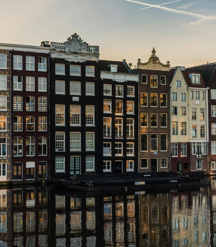 Rangée de maisons hautes et étroites avec de grandes fenêtres se reflétant dans l'eau au coucher du soleil à Amsterdam, Pays-Bas. Les bâtiments sont serrés les uns contre les autres et présentent des couleurs et des styles architecturaux variés.
