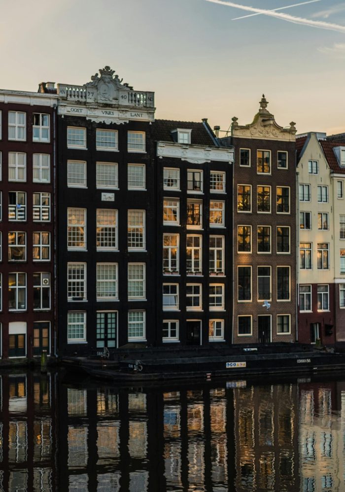 Rangée de maisons hautes et étroites avec de grandes fenêtres se reflétant dans l'eau au coucher du soleil à Amsterdam, Pays-Bas. Les bâtiments sont serrés les uns contre les autres et présentent des couleurs et des styles architecturaux variés.