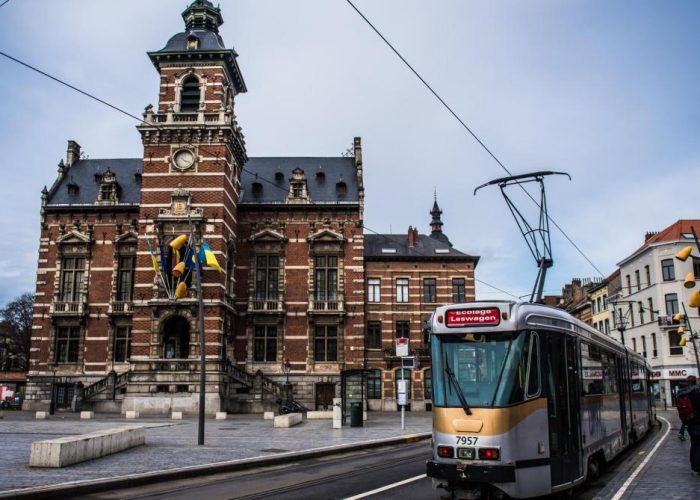 Un tramway moderne passe devant un bâtiment historique en briques avec une tour d'horloge sur une place de la ville, avec des drapeaux belges et des câbles de tramway visibles dans un ciel nuageux.