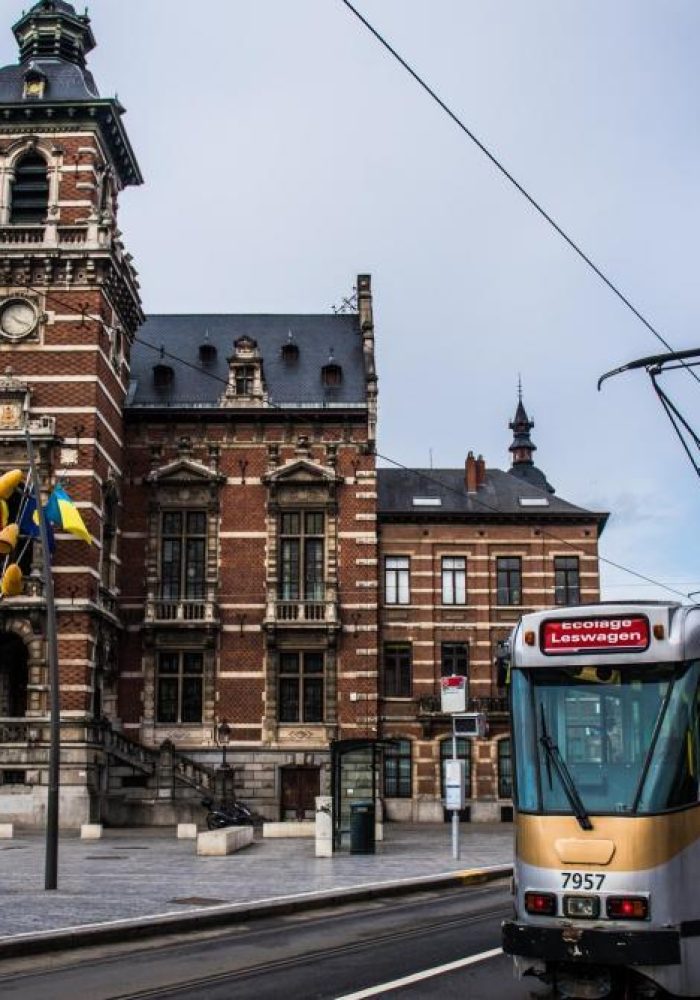 Un tramway moderne passe devant un bâtiment historique en briques avec une tour d'horloge sur une place de la ville, avec des drapeaux belges et des câbles de tramway visibles dans un ciel nuageux.