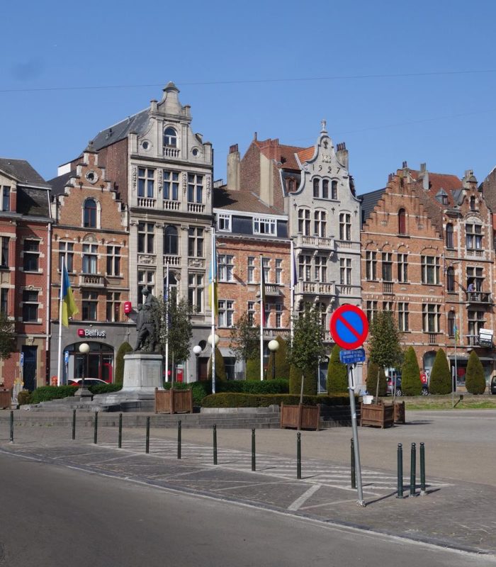 Une rangée de bâtiments historiques en briques ornés et coiffés de toits à pignons surplombe une place tranquille et ouverte par une journée ensoleillée. Un panneau d'interdiction de stationner et de petits arbres bordent la rue pavée au premier plan.